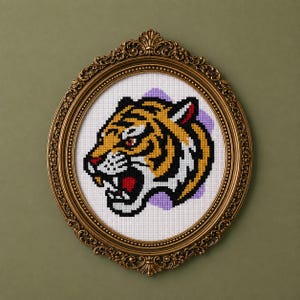 Könnte beinhalten: Eine Kreuzstichstickerei eines Tigerkopfes im Profil, gearbeitet mit gelbem, schwarzem und weißem Garn auf weißem Rasterstoff vor einem hellvioletten Hintergrund. Das Werk befindet sich in einem verzierten, ovalen Goldrahmen.