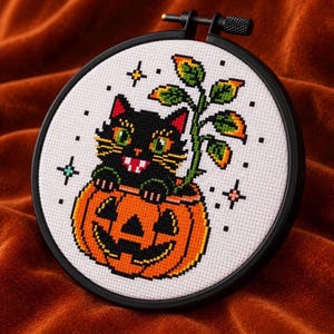 以下が含まれることがあります： 黒い刺繍枠の中に、緑色の目と白い牙を持つ黒猫がオレンジ色のジャック・オー・ランタンの中に入っているクロスステッチのデザイン。カボチャからはオレンジ色の先端を持つ緑色の植物が生えており、周囲には小さな星が散りばめられている。