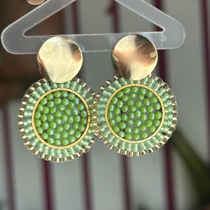 May include: A pair of round earrings with a gold-colored disc top and a green beaded center. The earrings have a gold-colored border and are held by a clear plastic hanger. The earrings are a bright, eye-catching accessory.