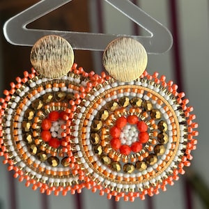 May include: A pair of handcrafted, beaded earrings. Each earring features a gold-colored, textured disc at the top, with a circular design of orange, white, and gold beads. The earrings are held by a clear hanger.