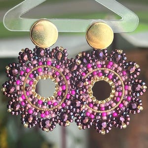 May include: A pair of handcrafted earrings featuring a gold-colored stud and a circular design. The earrings are adorned with a combination of small, dark purple, pink, and gold beads, creating a floral or sunburst pattern. The earrings are designed to dangle.