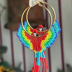 May include: Gold hoop earrings with a beaded parrot design. The parrot features red, yellow, green, and blue beads, with a red beaded tail. The earrings are suspended from a clear plastic hook.