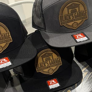 Op de afbeelding: Drie zwarte en grijze baseballpetten met een bruin leren patch waarop "Black's Garage Storage & Doors LLC" staat en een rood logo met de tekst "Flat Bill Snapback".