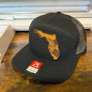 Könnte beinhalten: Eine schwarze und braune Trucker-Kappe mit flachem Schirm. Die Kappe hat einen braunen Leder-Patch mit dem Bundesstaat Florida und dem Staatssiegel. Die Kappe hat ein schwarzes Netzrücken und ein rotes "R"-Logo auf der Vorderseite.