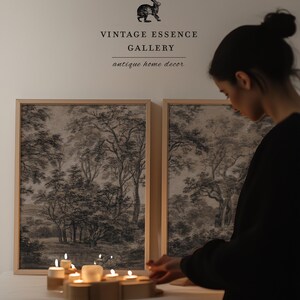 May include: Two framed black and white landscape prints of trees and a forest scene. A person is lighting candles on a table in front of the prints. The text above the prints reads "Vintage Essence Gallery antique home decor".