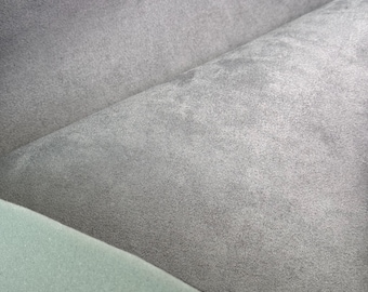 May include: Close-up of a gray fabric with a soft, velvety texture. The fabric is folded over, revealing a lighter gray underside and a section of light green foam. The image highlights the texture and color variations of the materials.