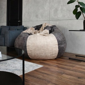 May include: A large, round beanbag chair in a modern living room. The beanbag has a colour-blocked design with grey, beige, and dark brown sections. The room also includes a dark blue sofa, a black side table, and a coffee table.
