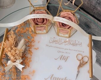 May include: A white tray with gold accents and a floral design. Two hexagonal glass boxes with gold frames and lids contain pink roses. The tray also includes a pair of gold scissors, a small bouquet of dried flowers, and a handwritten inscription that reads "He placed between you love and merry Quran 30:21 Alif & Anusha August 23, 2024".