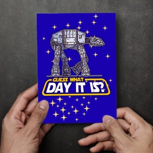 May include: A blue greeting card featuring a cartoon AT-AT walker illustration with the text "GUESS WHAT DAY IT IS?" in a yellow and white design, surrounded by yellow stars.