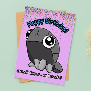 May include: A birthday card with a cartoon creature illustration. The card has the text "Happy Birthday!" in blue and "I smell danger... and snacks!" in black. The background is light purple with colorful confetti.