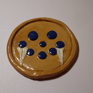 May include: A round, tan-colored clay coaster with a design of five blue blueberries and two white swirls.