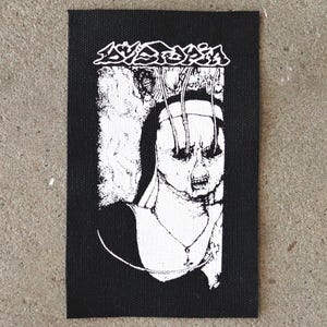 May include: Black and white patch with the word "Dystopia" above an illustration of a nun with wires connected to her eyes. The nun's face is distorted with an open mouth. The patch is rectangular.