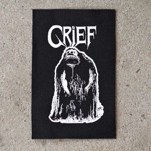 Pode incluir: Patch retangular preto com a palavra "GRIEF" em letras góticas brancas acima de uma ilustração branca de um monstro com a boca aberta. O patch está sobre uma superfície de concreto cinza.