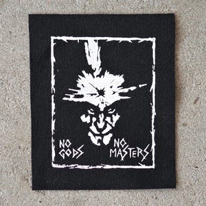 May include: Black square patch with a white graphic of a stylized face within a white rectangular border. The text "NO GODS NO MASTERS" is printed in white on the patch.