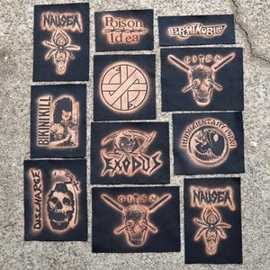 Op de afbeelding: Een verzameling zwarte stoffen patches met verschillende bandlogo's en afbeeldingen in een verbrand oranje. De patches bevatten tekst zoals "NAUSEA", "Poison Idea", "Bikini Kill" en "Exodus", samen met schedel- en spinnenillustraties.