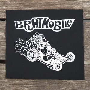 May include: A black iron-on patch with a white illustration of a cartoon character driving a race car with the text "Bratmobile" above it.