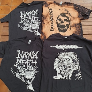 Puede incluir: Cuatro camisetas negras con diseños gráficos blancos. Los diseños incluyen los nombres de las bandas "Napalm Death" y "Discharge" e imágenes gráficas de cráneos, una mano sosteniendo un cuenco y una cara gritando.