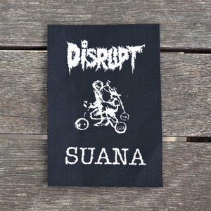Può includere: Toppa ricamata in bianco e nero con il testo "DISRUPT SUANA" in carattere gotico. La toppa presenta una grafica di due figure che guidano una motocicletta.