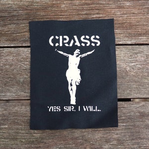 以下が含まれることがあります： 白と黒のアイロン接着パッチで、バンド名「CRASS」が白いステンシル文字で書かれています。パッチの中央には、腕を広げた人の白いシルエットがあります。パッチの下部には「YES SIR, I WILL...」というテキストがあります。