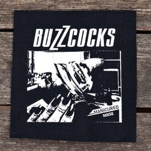 Peut inclure: Écusson carré noir avec le nom du groupe "BUZZCOCKS" en blanc. Un graphisme noir et blanc montre une main aux longs ongles tenant un appareil. Le texte "MANICURED NOISE" est en dessous, dans un style punk rock.
