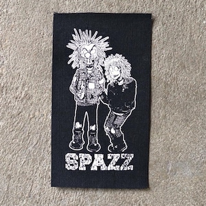 Op de afbeelding: Zwart rechthoekig embleem met een witte illustratie van twee stripfiguren met stekelig haar en kleding. Het woord "SPAZZ" is in witte blokletters onderaan gedrukt. De patch ligt op een neutraal gekleurd oppervlak.