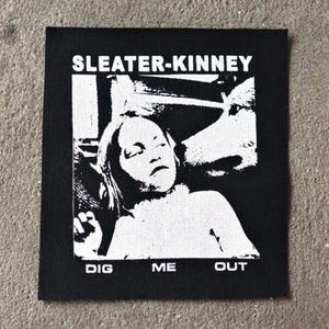 Puede incluir: Parche cuadrado en blanco y negro con el texto "SLEATER-KINNEY" sobre una imagen de una persona y un perro. Las palabras "DIG ME OUT" están impresas debajo. El parche es cuadrado.