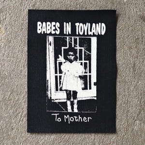 May include: Black fabric patch with the text "BABES IN TOYLAND" above a black and white image of a girl in a doorway. The text "To Mother" is below the image. The patch is rectangular.