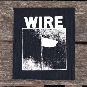 Op de afbeelding: Zwart vierkantige patch met het woord "WIRE" in witte letters boven een witte afbeelding van een vlag. De patch ligt op een houten oppervlak.