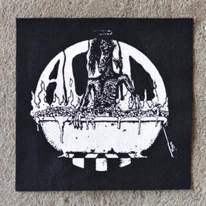 K&ouml;nnte beinhalten: Schwarz-wei&szlig;er Aufn&auml;her mit einer verst&ouml;renden Illustration einer Figur in einer Wanne, umgeben von Flammen. Die Buchstaben "AC" sind in einem Kreis zu sehen. Der Aufn&auml;her ist quadratisch und aus Stoff.