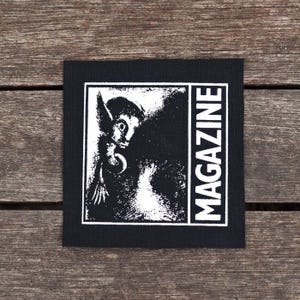 May include: Black square patch with a white illustration of a creature with large ears and the word "MAGAZINE" in white vertical text. The patch is on a wooden surface.