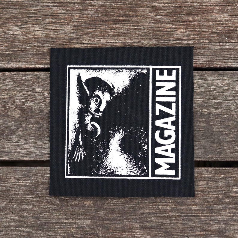 May include: Black square patch with a white illustration of a creature with large ears and the word "MAGAZINE" in white vertical text. The patch is on a wooden surface.
