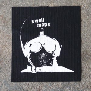 May include: Black square patch with a white graphic of a figure wearing goggles and a mask. The text "swell maps" is above the figure's head in white letters. The patch is made of a woven fabric.