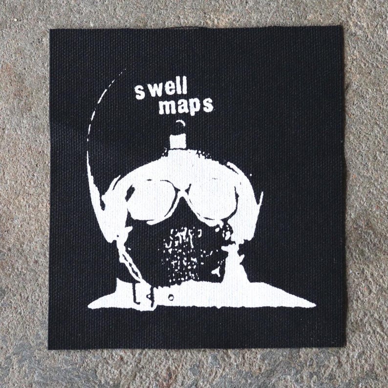 May include: Black square patch with a white graphic of a figure wearing goggles and a mask. The text "swell maps" is above the figure's head in white letters. The patch is made of a woven fabric.