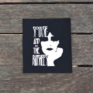 May include: Black square patch featuring a white graphic of a face and the text "Siouxsie and the Banshees" in a stylized font. The patch is set against a wooden background.