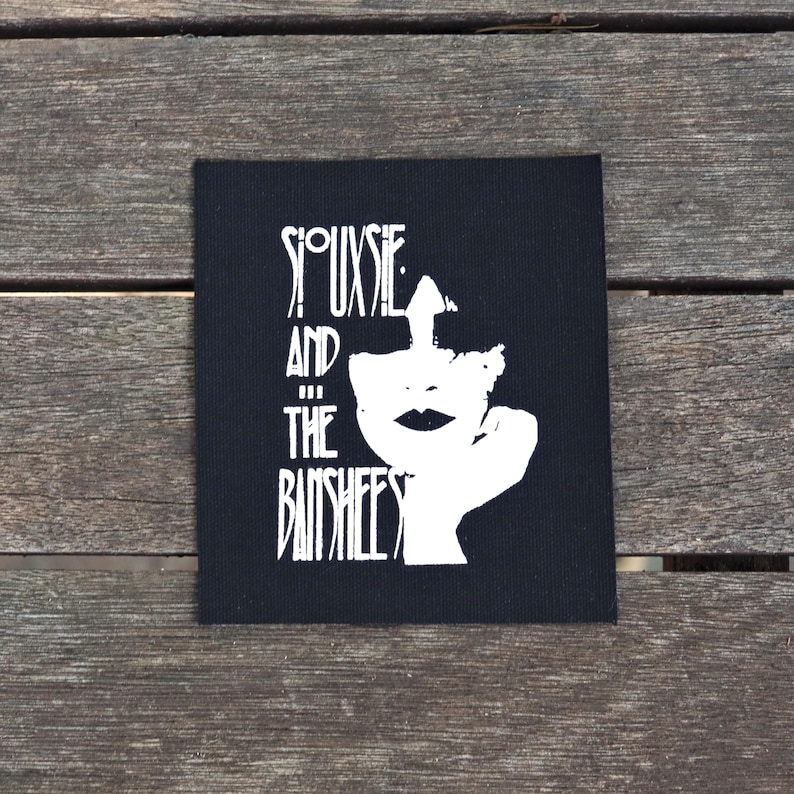 May include: Black square patch featuring a white graphic of a face and the text "Siouxsie and the Banshees" in a stylized font. The patch is set against a wooden background.