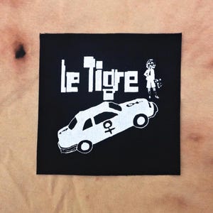Puede incluir: Parche bordado en blanco y negro con el texto "Le Tigre" y una imagen de dibujos animados de un coche y una persona.