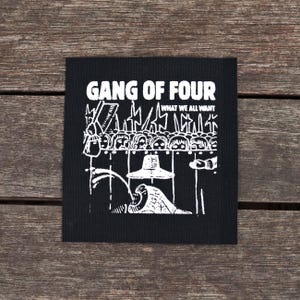 May include: Black square patch with white graphic art and text. The text reads "GANG OF FOUR" above "WHAT WE ALL WANT". The graphic depicts a crowd of faces, a hat, and other abstract shapes. The patch is on a wooden surface.