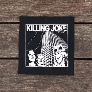 May include: Black square patch with the text "KILLING JOKE" above a white illustration of a skyscraper, lightning, and faces. The patch is likely for a jacket or bag, and is made of fabric.