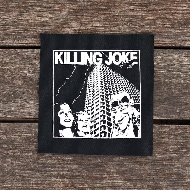 May include: Black square patch with the text "KILLING JOKE" above a white illustration of a skyscraper, lightning, and faces. The patch is likely for a jacket or bag, and is made of fabric.