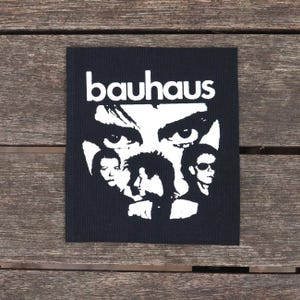 May include: Black square patch featuring a white graphic of the band Bauhaus. The design includes the band's name and stylized portraits of the band members. The patch is on a wooden surface.