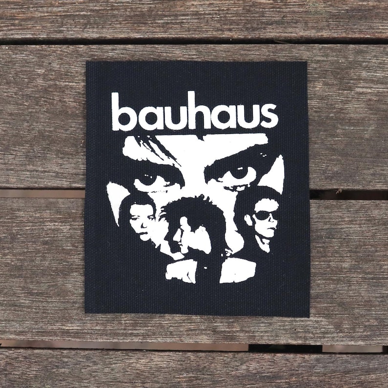 May include: Black square patch featuring a white graphic of the band Bauhaus. The design includes the band's name and stylized portraits of the band members. The patch is on a wooden surface.