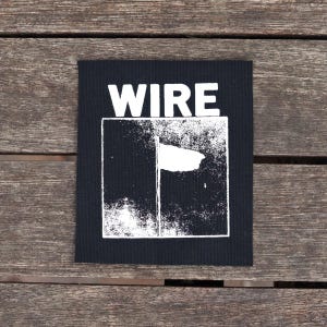 May include: Black square patch with the word "WIRE" in bold white letters at the top. Below, a white graphic design is set within a white-lined rectangle. The patch is on a wooden surface.