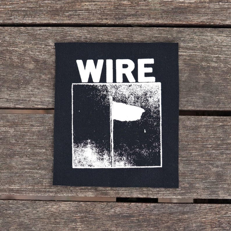 May include: Black square patch with the word "WIRE" in bold white letters at the top. Below, a white graphic design is set within a white-lined rectangle. The patch is on a wooden surface.