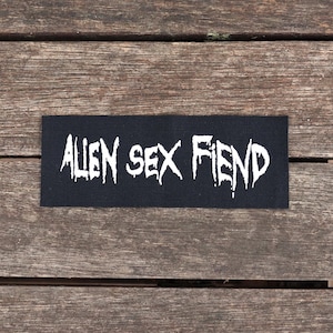 May include: A black rectangular patch with the words "ALIEN SEX FIEND" in white, stylized, jagged lettering. The patch is set against a weathered wooden surface.