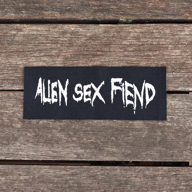 May include: A black rectangular patch with the words "ALIEN SEX FIEND" in white, stylized, jagged lettering. The patch is set against a weathered wooden surface.