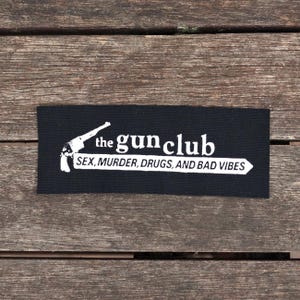 May include: A black rectangular patch with white text and a pistol graphic. The text reads "the gun club" above "SEX, MURDER, DRUGS, AND BAD VIBES" in a white arrow.
