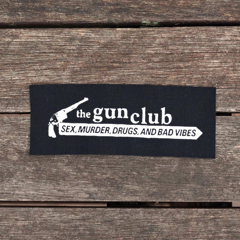 May include: A black rectangular patch with white text and a pistol graphic. The text reads "the gun club" above "SEX, MURDER, DRUGS, AND BAD VIBES" in a white arrow.