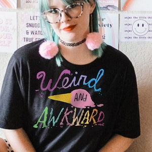 May include: A woman wearing a black t-shirt with the text "Weird and Awkward" in rainbow colors. She is sitting on a white stool with her legs crossed. She has green hair and is wearing glasses. The background is a wall with a variety of colorful prints.