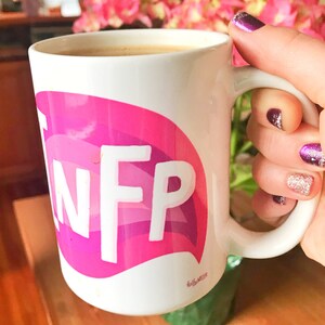 May include: White ceramic mug with a pink and purple speech bubble design that says "INFP". The mug is filled with coffee and is being held by a hand with purple nail polish.