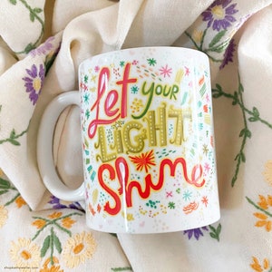 Puede incluir: Taza de cer&aacute;mica blanca con las palabras "Let Your Light Shine" en letras coloridas. La taza tiene un asa blanca y est&aacute; decorada con varias formas y dise&ntilde;os coloridos. La taza est&aacute; sobre un pa&ntilde;o color crema con flores bordadas.
